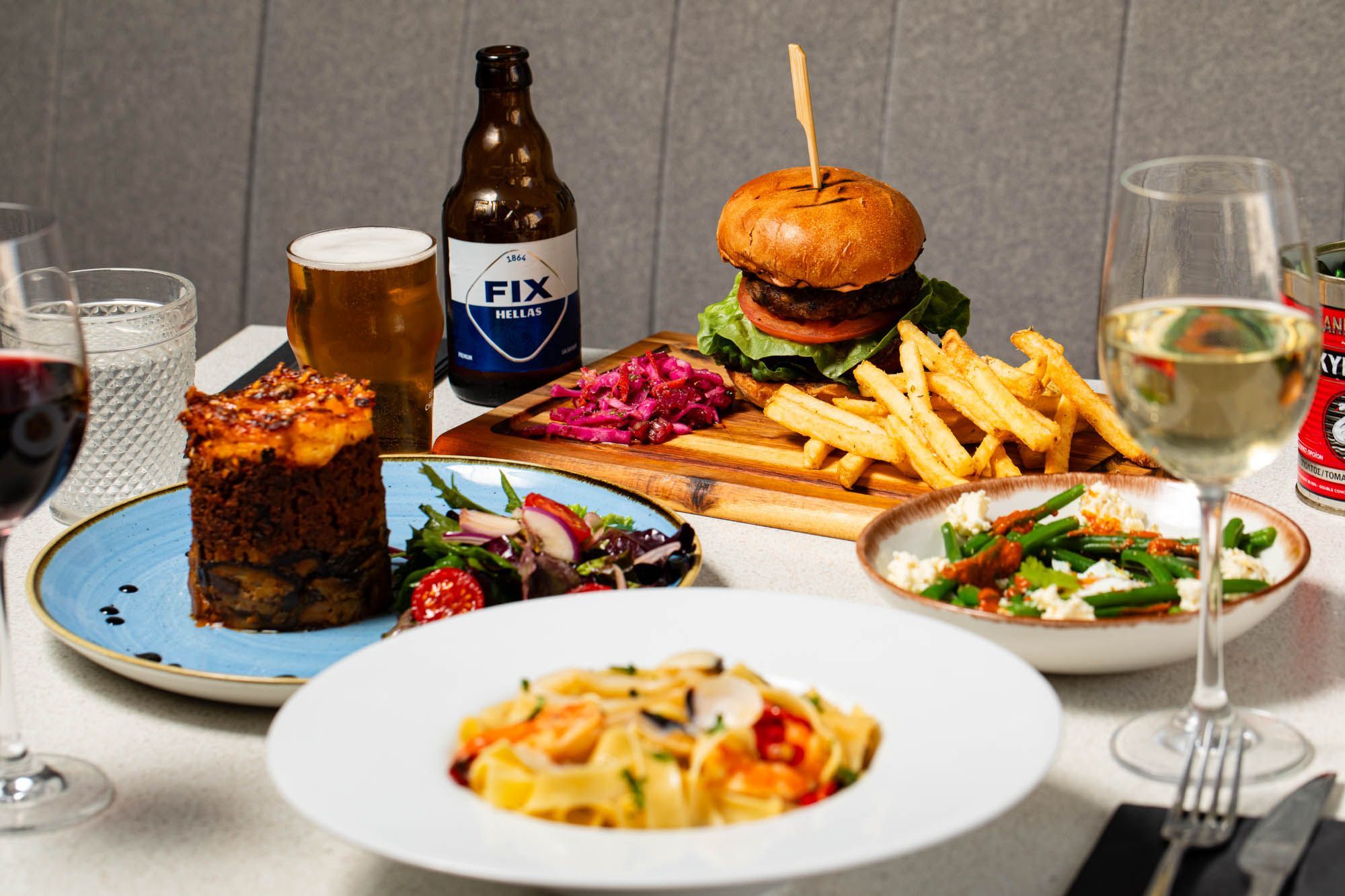 side shot of the table laid out with few dishes inlsuding burger and chips, pasta, drinks