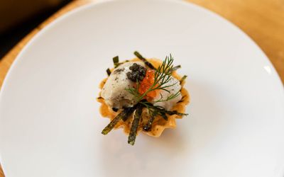 A white circular plate rests on the edge of a circular wooden table. At the centre of the plate is a small stack of food, red and black caviar, samphire a foam reduction