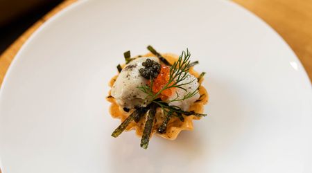 A white circular plate rests on the edge of a circular wooden table. At the centre of the plate is a small stack of food, red and black caviar, samphire a foam reduction