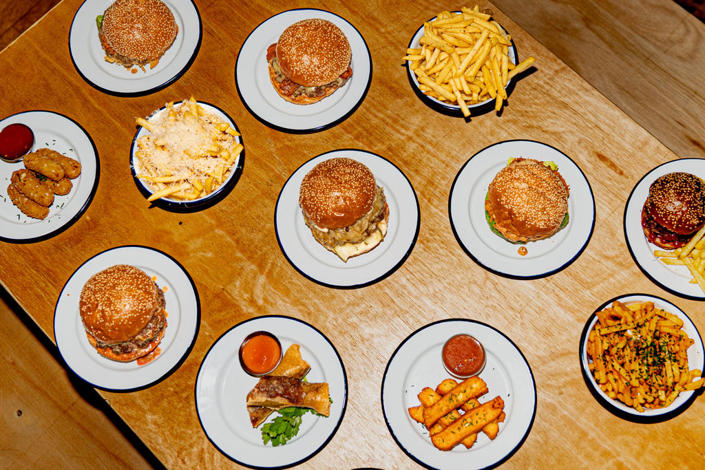 over head shot of the light brown wooden table laid out with bowls of chips and multiple burgers. Shelter Hall is a food market on Brighton's seafront. A restaurant with many different food types to choose from and a Brighton burger restaurant is one of the options.