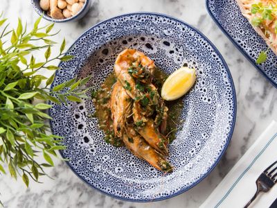 Whole prawns in a garlic sauce with a wedge of lemon served in a blue ceramic plate with a leafy plant displayed next to the plate.