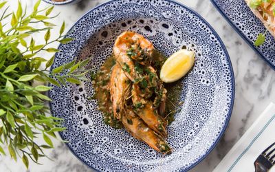 Whole prawns in a garlic sauce with a wedge of lemon served in a blue ceramic plate with a leafy plant displayed next to the plate.