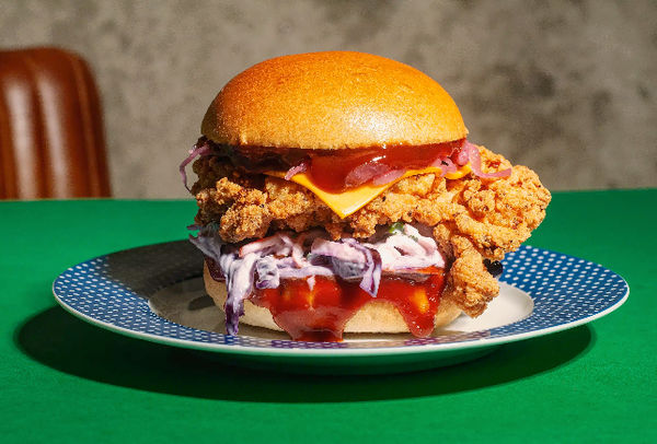 A chicken burger stacked with fried chicken in a bun with slaw and sauce, served on a plate on a green table.