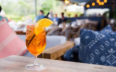 A glass of Aperol Spritz with a garnish for fresh orange sat on a table with a restaurant view in the background.