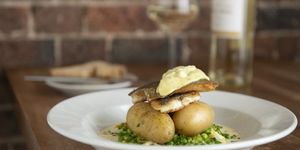A large shallow dish with a bed of peas topped with new potatoes, a piece of fish and butter. Photographed with a bottle of white wine next to a brick wall.