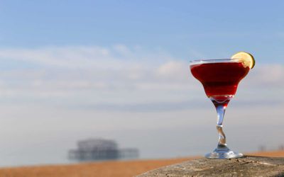 A bright red cocktail in a glass with a wobbly stem garnished with a wedge of lime. Photographed on the beach with the West Pier in the background.