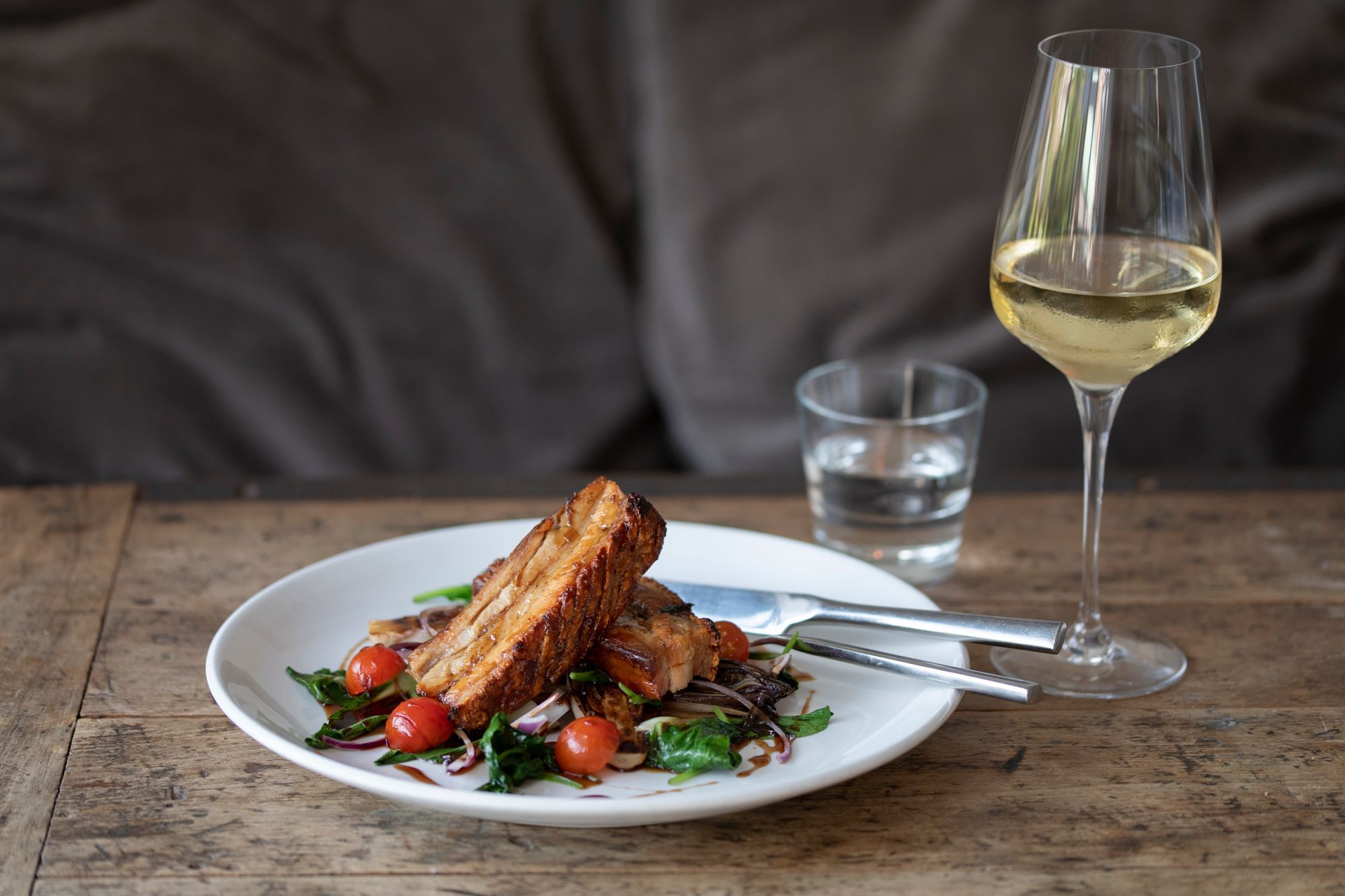 Salmon on top of colourful salad and 3 cherry tomatoes. the dish is on a white plate on a solid wood table with glass of white wine in the background. Morleys Bistro Hurstpierpoint