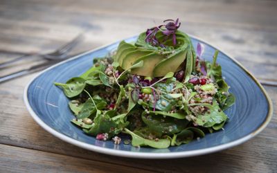 A leafy salad with spinach, sprouts and thinly sliced avocado. Served on a blue triangular plate on a wooden table.