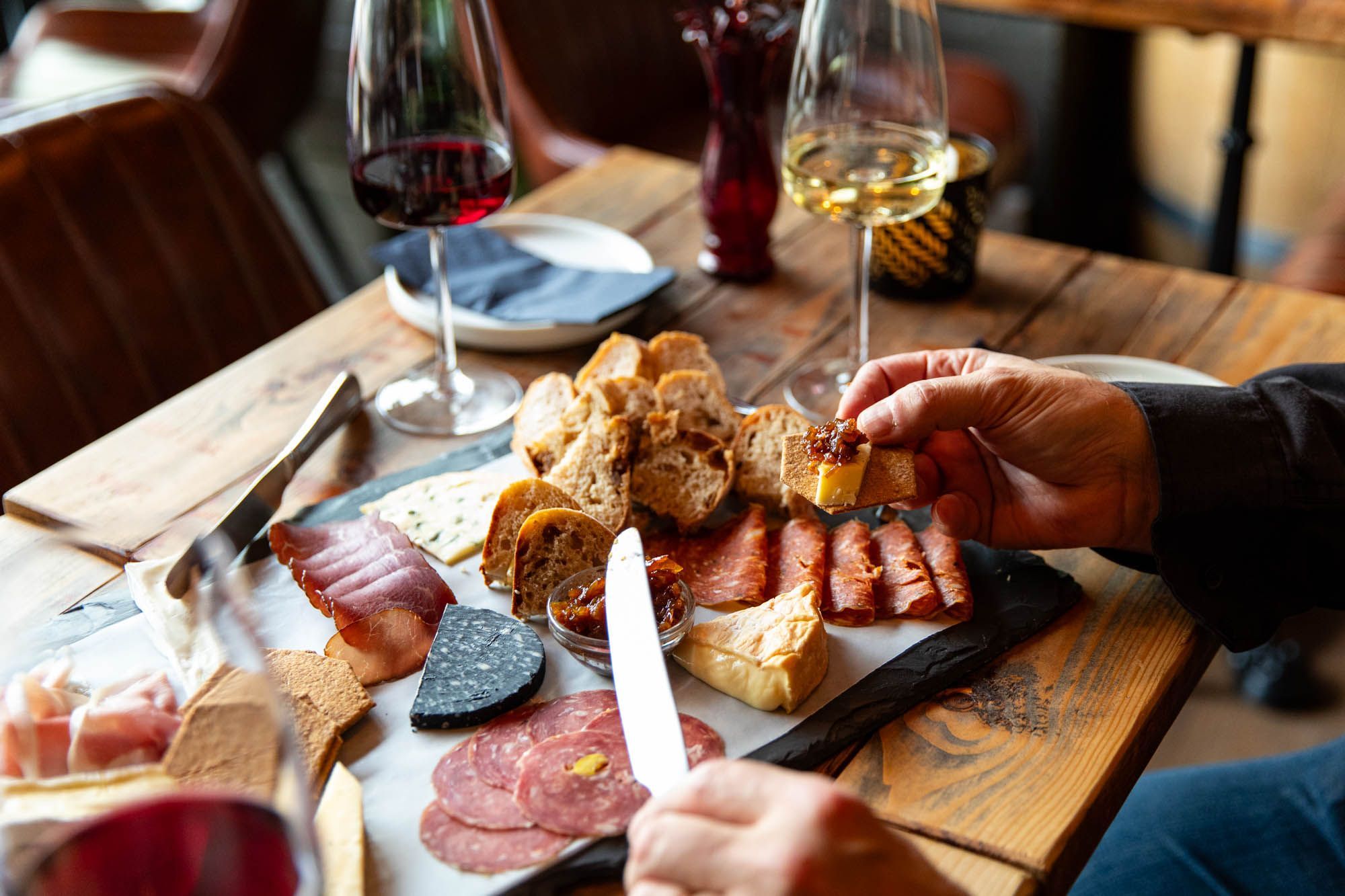 person spreading chutney on bread with piece of cheese, charcuterie board next to it and glass of both red and white wine