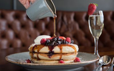A stack of pancakes with blueberries and a jug of syrup being poured over them. Served with a glass of champagne.
