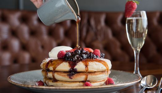 A stack of pancakes with blueberries and a jug of syrup being poured over them. Served with a glass of champagne.