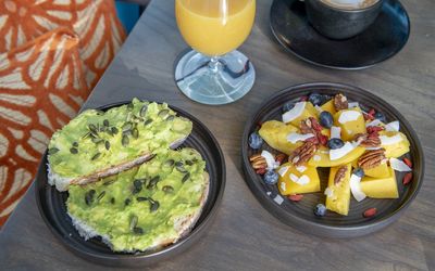 Smashed avocado on toast with a fruit salad and glass of juice at Cyan. Positioned on a faux wooden table. This venue is part of the Brighton Breakfast Guide.