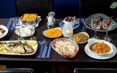 A feast of Greek food displayed on plates, bowls including a whole fish, dips and sides. Served on a black table with dark interiors.
