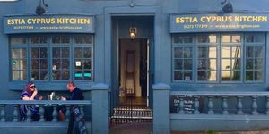 Photograph of the front street exterior and alfresco dining of Estia restaurant with dark blue paintwork, windows and gold signage.