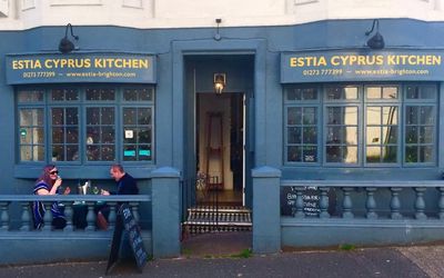 Photograph of the front street exterior and alfresco dining of Estia restaurant with dark blue paintwork, windows and gold signage. Greek Restaurant Brighton