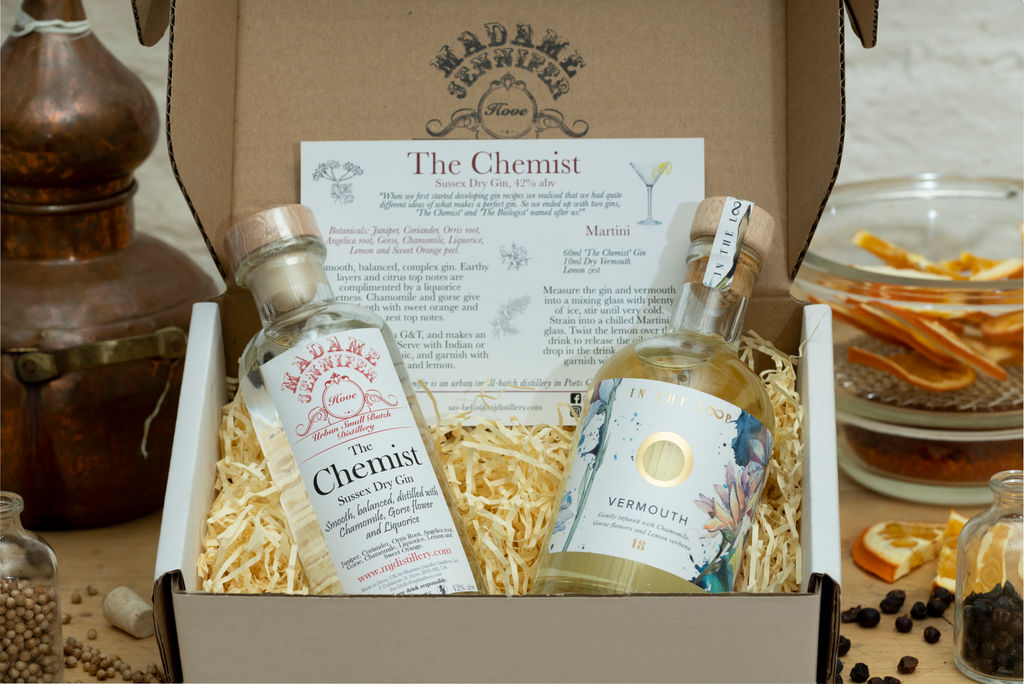 A cocktail mixing kit from Madame Jenifer's a bottle of gin and a bottle of vermouth and instructions for mixing a martnini. In the background there aer various flavourings in glass jars and bowls and an old fashioned copper still