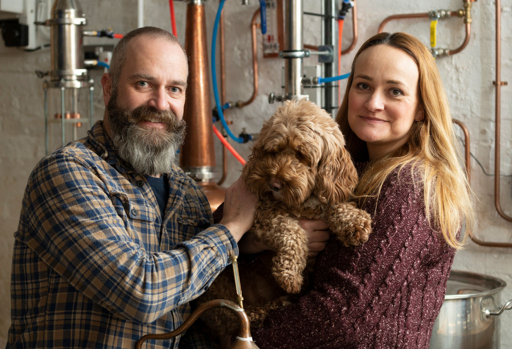 To the left a man in a plaid shirt, he has a thick beard and handlebar moustache, to the right there is a strawberry blond haired woman in a mauve jumper. They are holding a brown dog between them, in the background there are lots of pipes