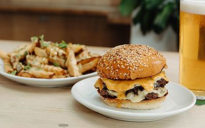 A small plate with a seeded bun hamburger and plate of wedges. Served with a pint of beer on a wooden dining table with a plant in the background. Brighton Beer Garden