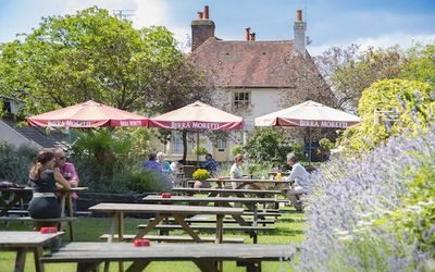 A country beer garden with wooden benches and parasols. A picturesque cottage in the background and lavender bushes.