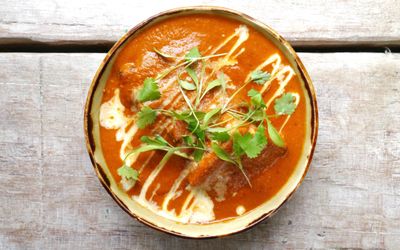 Overhead shot of a bowl filled with a bright orange, creamy tomato based curry garnished with herbs and drizzled with cream.