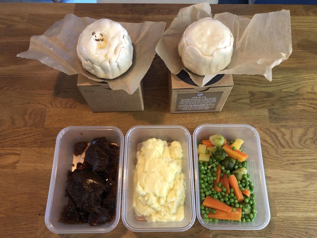 two uncooked pies on top of brown kraft boxes and three containers of vegetables, mashed potato and onion gravy laid out on a wooden table