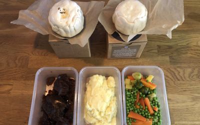 Two uncooked pies on top of brown kraft boxes and three containers of vegetables, mashed potato and onion gravy laid out on a wooden table