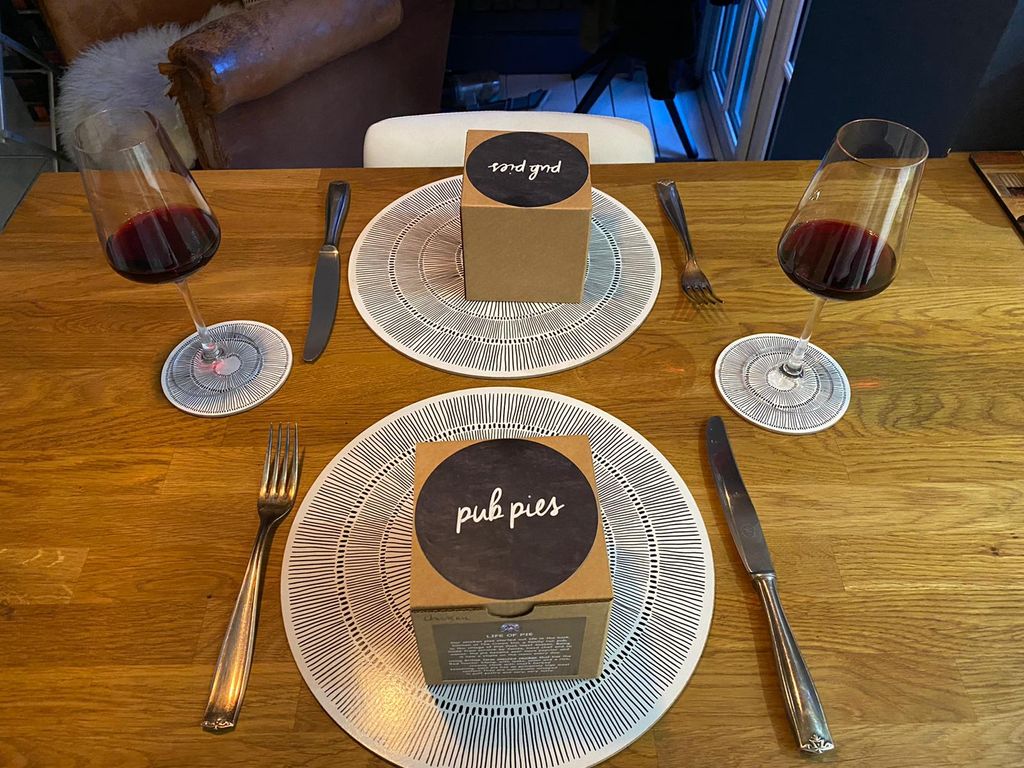 two boxes on plates at a dining table set for two with two glasses of wine. The brown boxes have black round labels on them that read "pub pies"