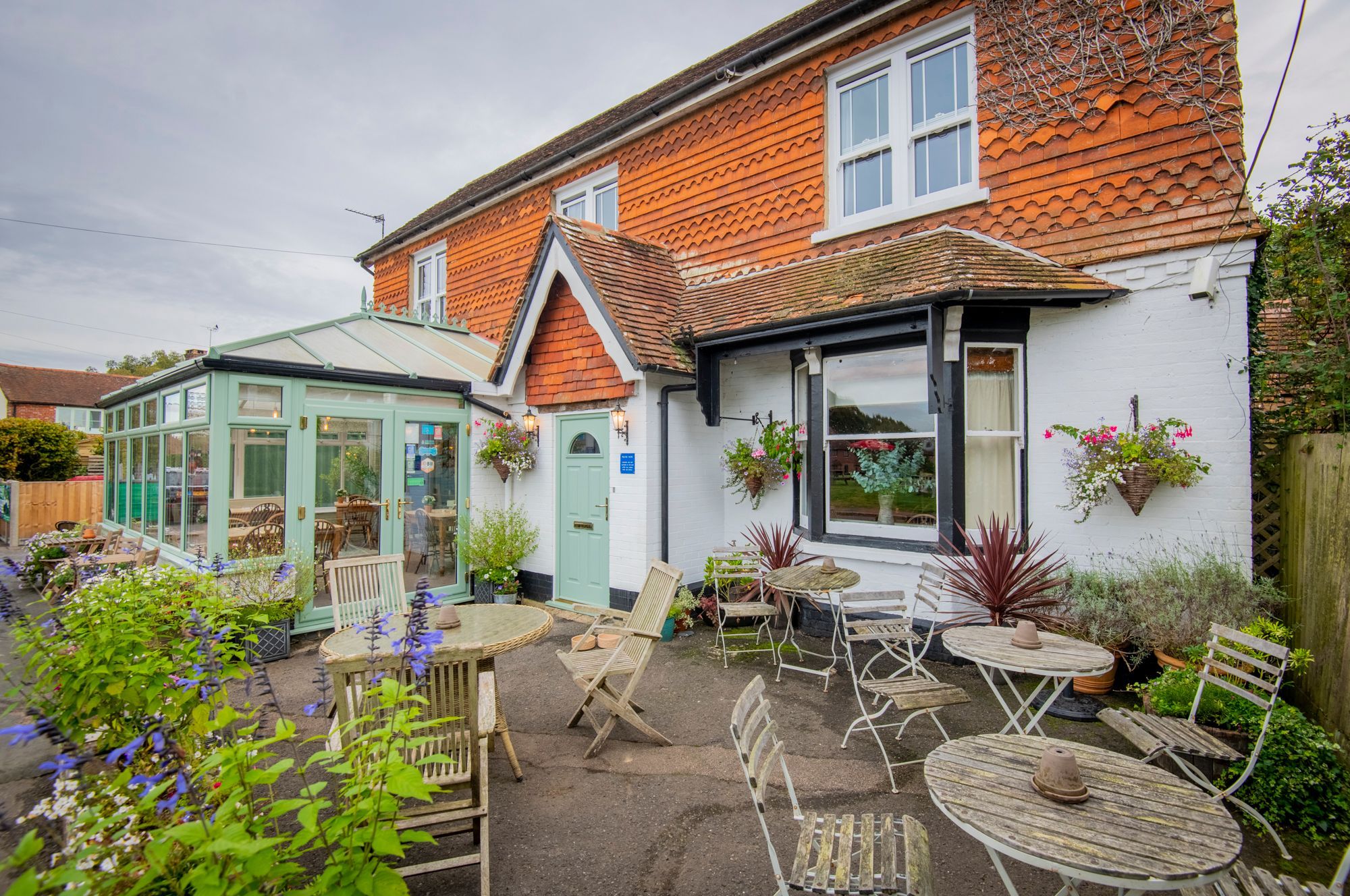 The front of The Crown Inn Dial Post pub with patio seating and a conservatory and a raised flower bed at the front. The pub is white washed with red tilled gables and roof and a sage green front door.