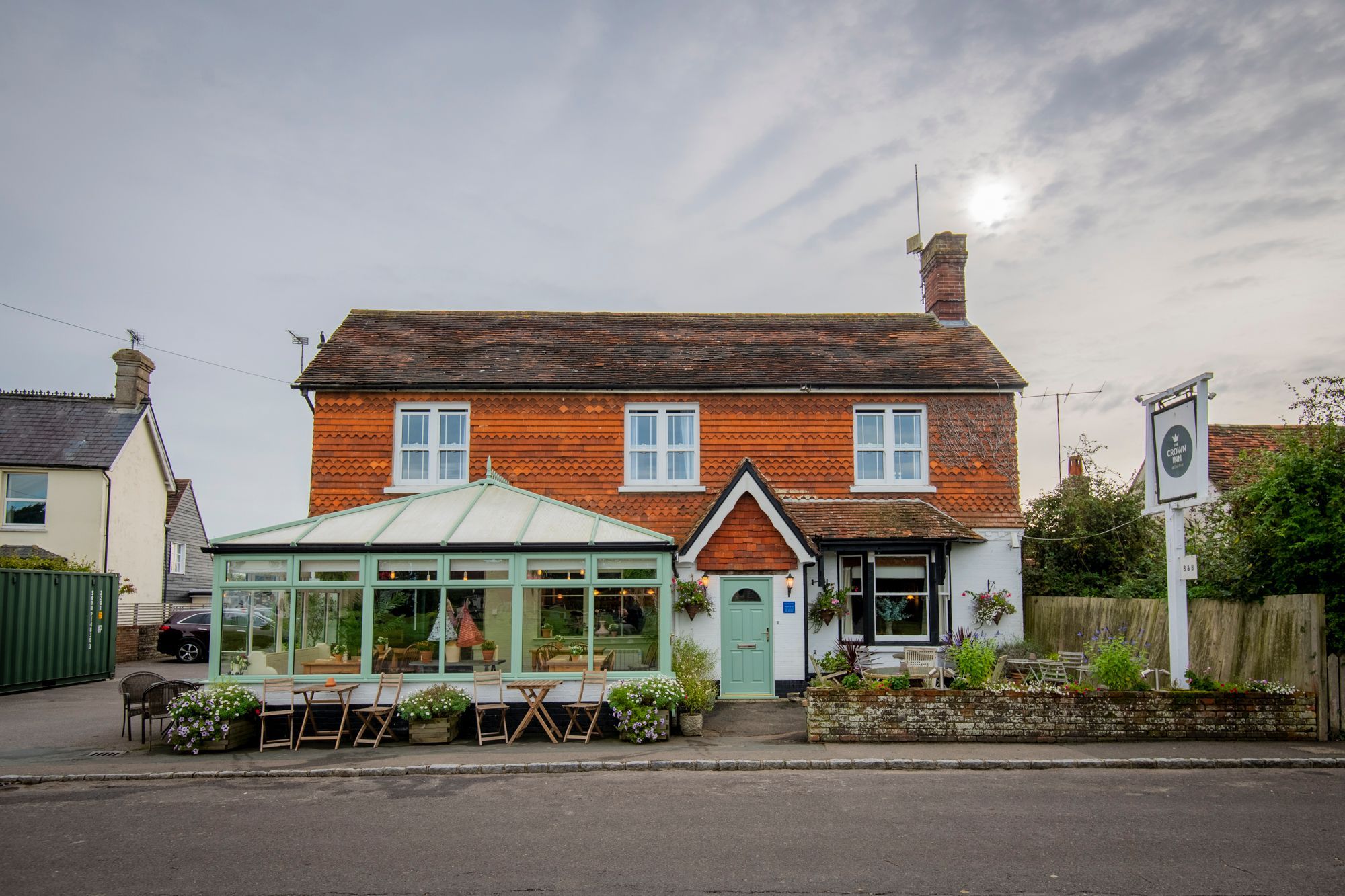 The front of The Crown Inn Dial Post pub with patio seating and a conservatory and a raised flower bed at the front. The pub is white washed with red tilled gables and roof and a sage green front door.