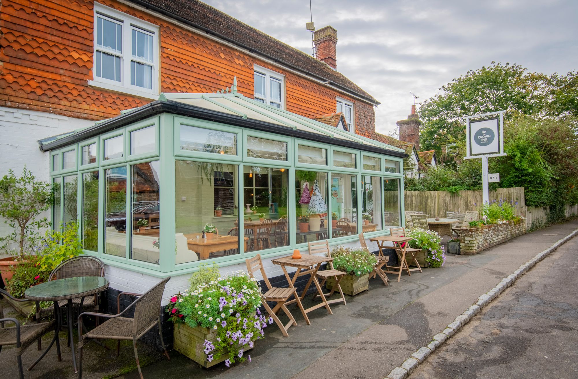 The front of The Crown Inn Dial Post pub with patio seating and a conservatory and a raised flower bed at the front. The pub is white washed with red tilled gables and roof and a sage green front door.