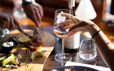 A hand holding a glass of rosé wine is opposite a pair of hands with cutlery taking food from a charcuterie board.