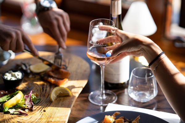 A hand holding a glass of rosé wine is opposite a pair of hands with cutlery taking food from a charcuterie board.