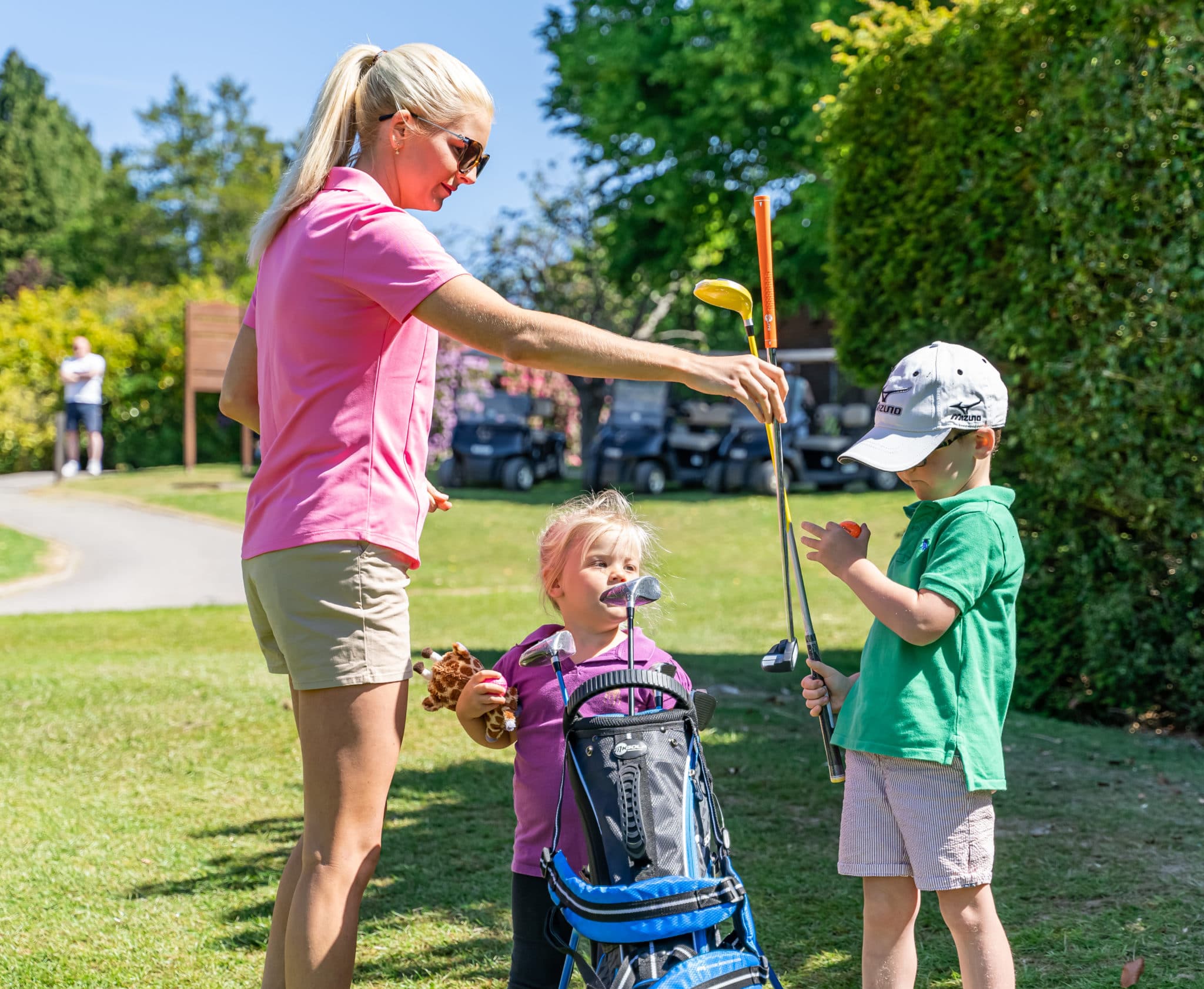 Two children are being shown how to play gold, a woman in a pink t-shirt is handing a club to a boy in a green t-shirt and cap, the second child is stood with the golf club bag.