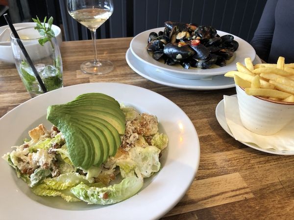 Plates of food laid out on a wooden table with a sliced avocado salad, moules mariniere and fries.