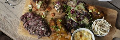 A wooden chopping board serving slice meat, salad, a pot of coleslaw and a pot of sauce with a glass of red wine.