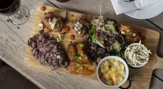A wooden chopping board serving slice meat, salad, a pot of coleslaw and a pot of sauce with a glass of red wine.