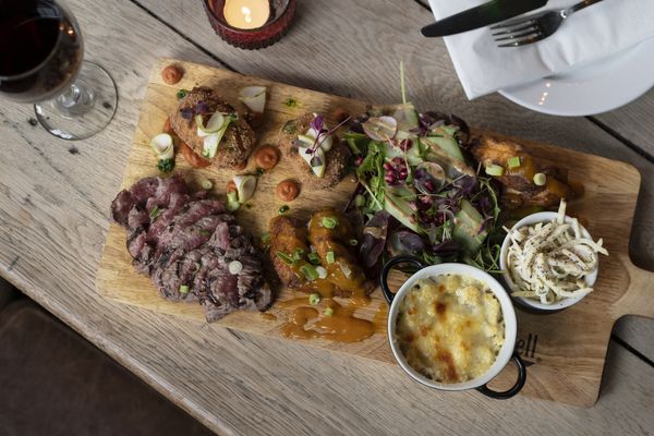 A wooden chopping board serving slice meat, salad, a pot of coleslaw and a pot of sauce with a glass of red wine.