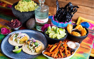Mexican feast presented featuring tacos, tequila, lime wedges, fries and dips.