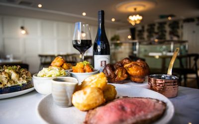 Roast beef and roast potatoes on a plate with a small pot of gravy, in the background there are bowls of vegetables, Yorkshire puddings and an open bottle of red wine with a glass full next to it.