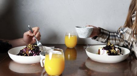 A table set with two brunch bowls coffee and orange juice, just in shot are two people sat opposite each other, the person on the right is raising a coffee cup, the person on the left is bringing a fork to their bowl.