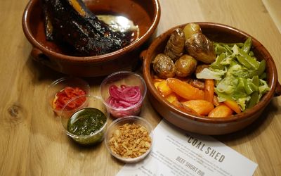 A bowl of sides including potatoes and carrots, a plate of chargrilled meat with dips and sundries. Presented with the Coal Shed paper menu.