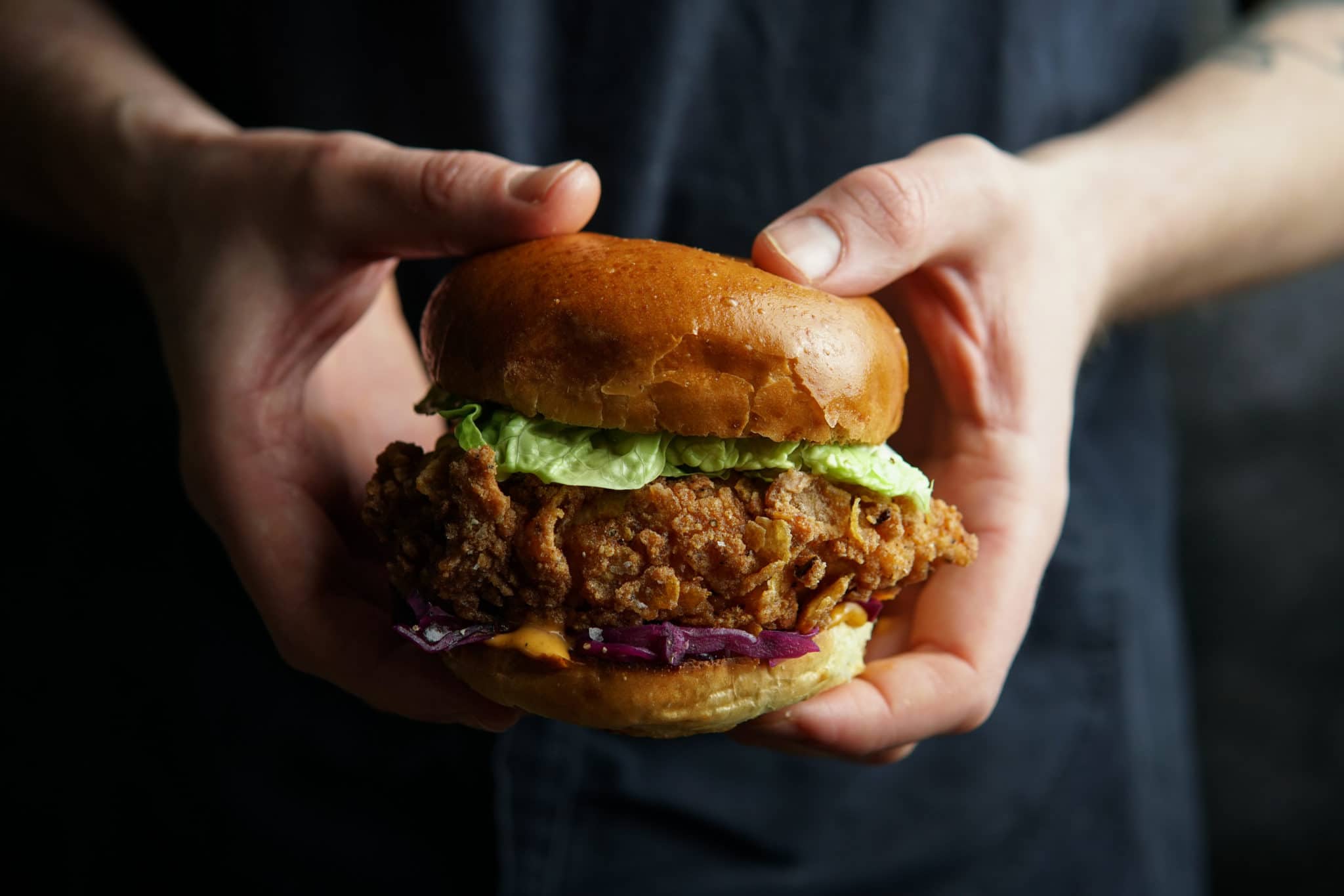 Hands holding a Southern fried fish burger with lettuce and red cabbage slaw.