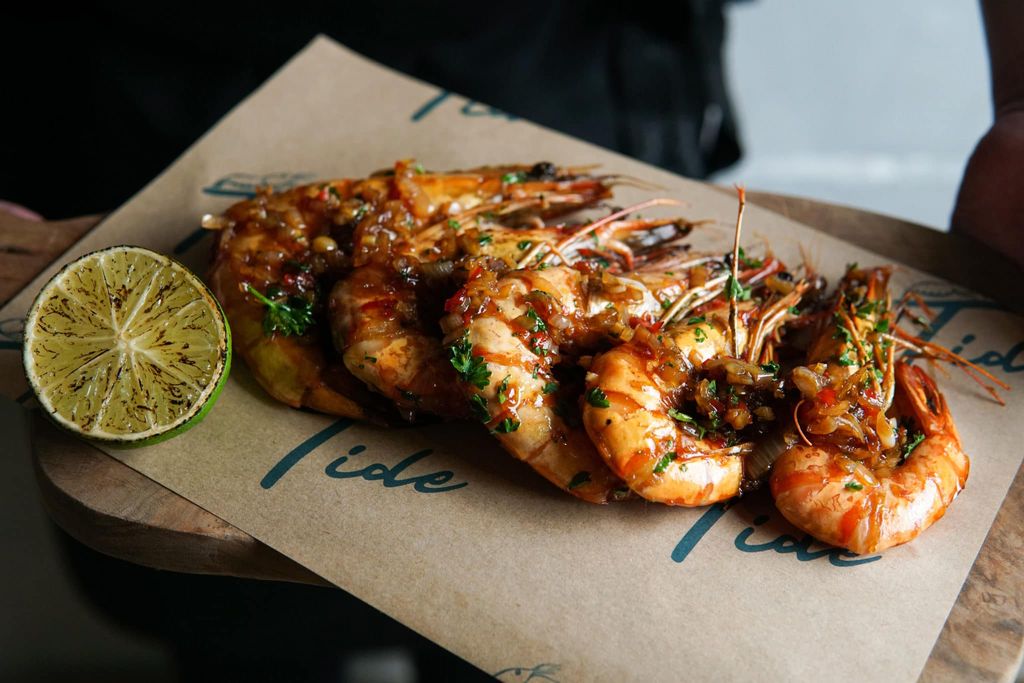 Rum tiger prawns with a burnt lime half, served on a wooden board with a piece of parchemnt on it and the word Tide in blue written on it.