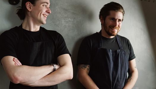 The Tide team, Chefs James and Raph. Raf to the left is earing a balck t-shirt and is looking over his left sholder at James. James, also in a black t-shirt and navy apron holds his hands in his lap and smiles while looking forward.