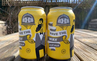 Yellow and grey artwork on two craft ale beer cans sat on a wooden table in the sun