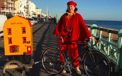 Lady dressed in red with a bicycle and house shaped trailer next to Brighton beach