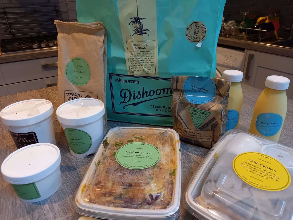 A blue paper bag with Dishoom written on it, white card food containers and biodegradable food containers with rice in them and bottles of drink