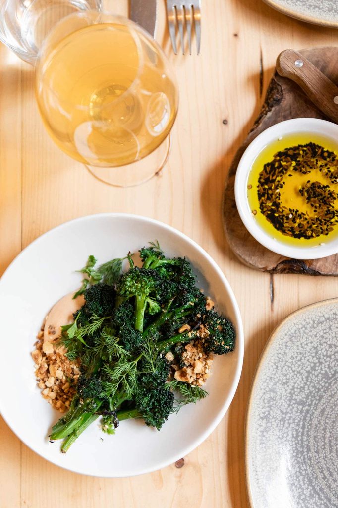 On overhead shot of a wooden table with a dish of broccoli, a vinaigrette and a glass of orange wine