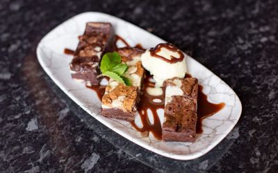 white plate with three chocolate brownies on covered with chocolate toppings and one scoop of vanilla ice cream