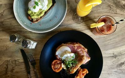 Bacon, egg and tomato alongside a plate of avocado on toast with a poached egg. Served with juice on a rustic wooden table.